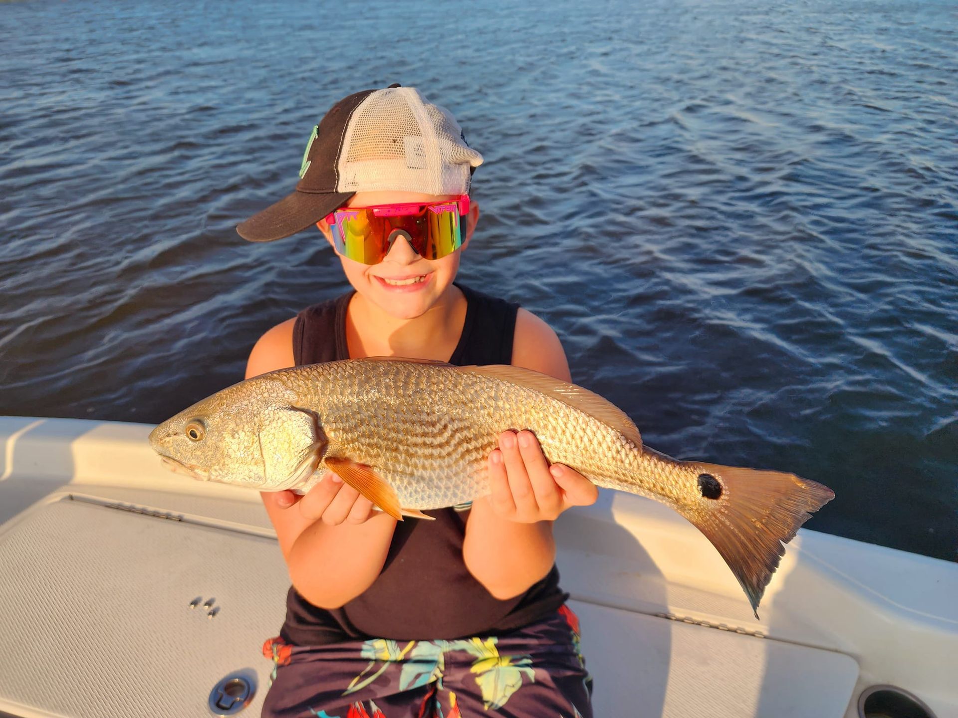 Boy smiling, holding a redfish on a boat, wearing sunglasses and hat, water in background.