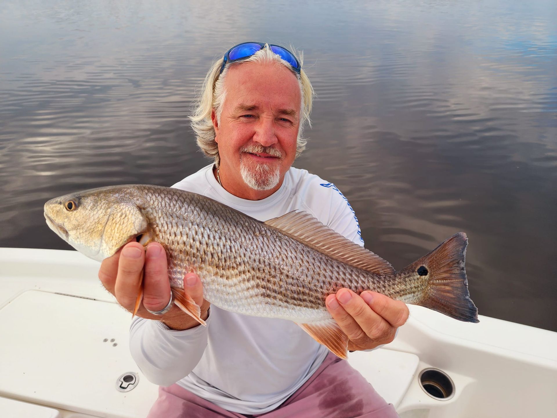 Man holding a redfish on a boat, smiling. Graying hair, sunglasses, on water.