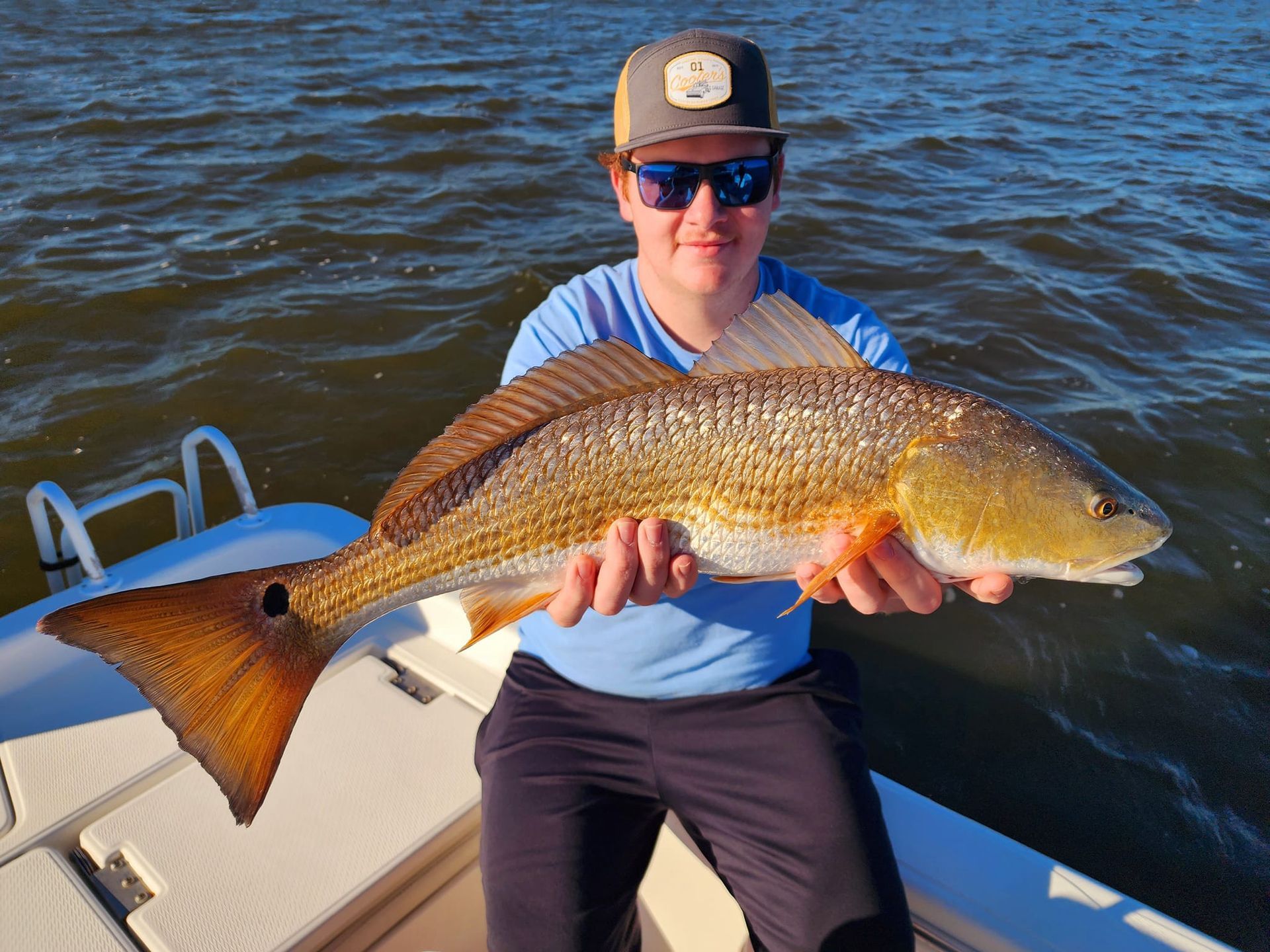 Man on boat holds a large redfish. Brown water and sunny sky in the background.