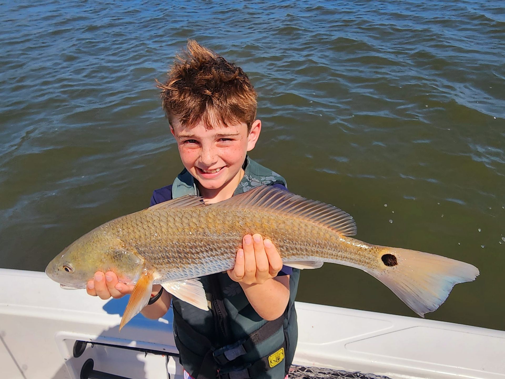 Boy holding a large redfish on a boat, smiling, blue water background.