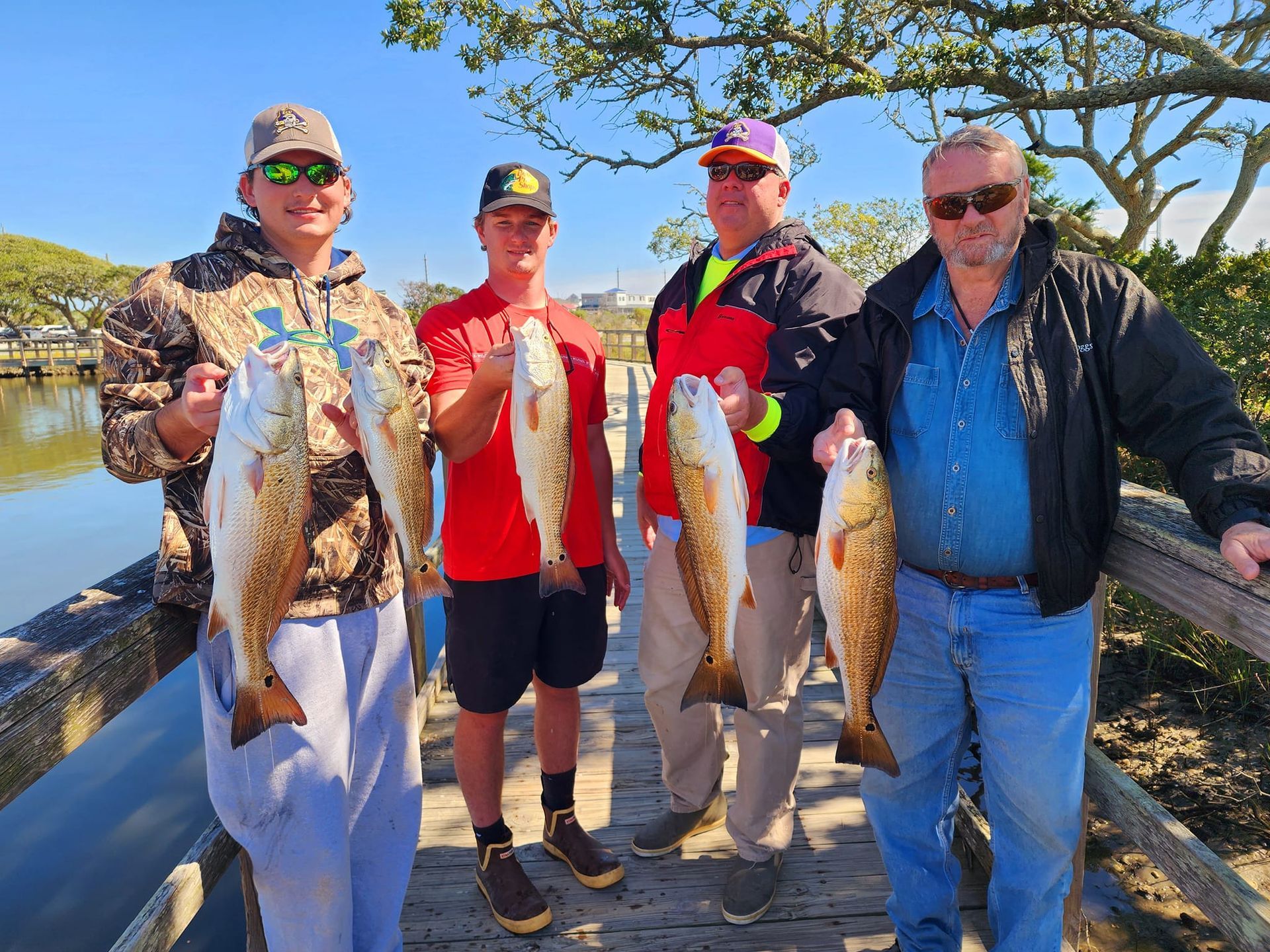 Four people holding up large redfish on a pier. Sunny day, trees in background, water on left.