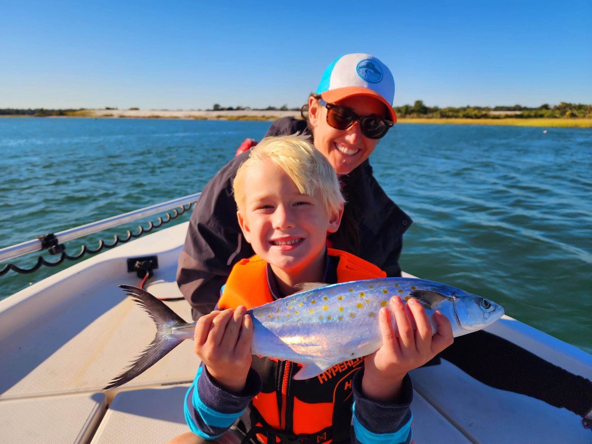 Boy smiles, holding fish on boat with person. Outdoors, sunny day on water.