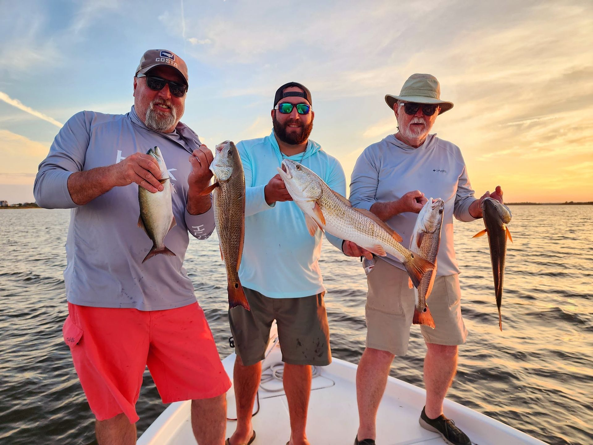 Three people on a boat display their caught fish at sunset; two redfish and two others.