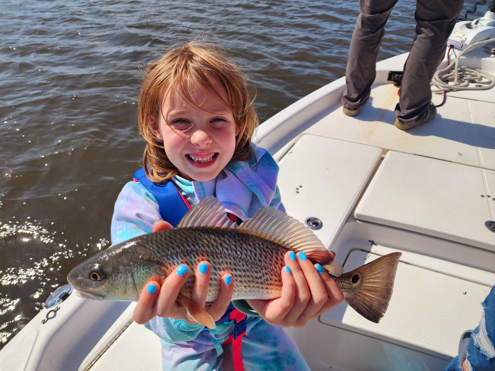 Young girl smiles, holding a redfish on a boat, blue nails, water in background.