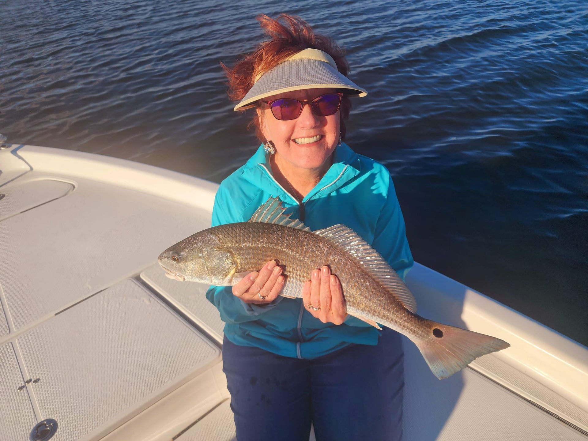 Woman on a boat smiles, holding a redfish. Sunlight on water, blue jacket, visor.