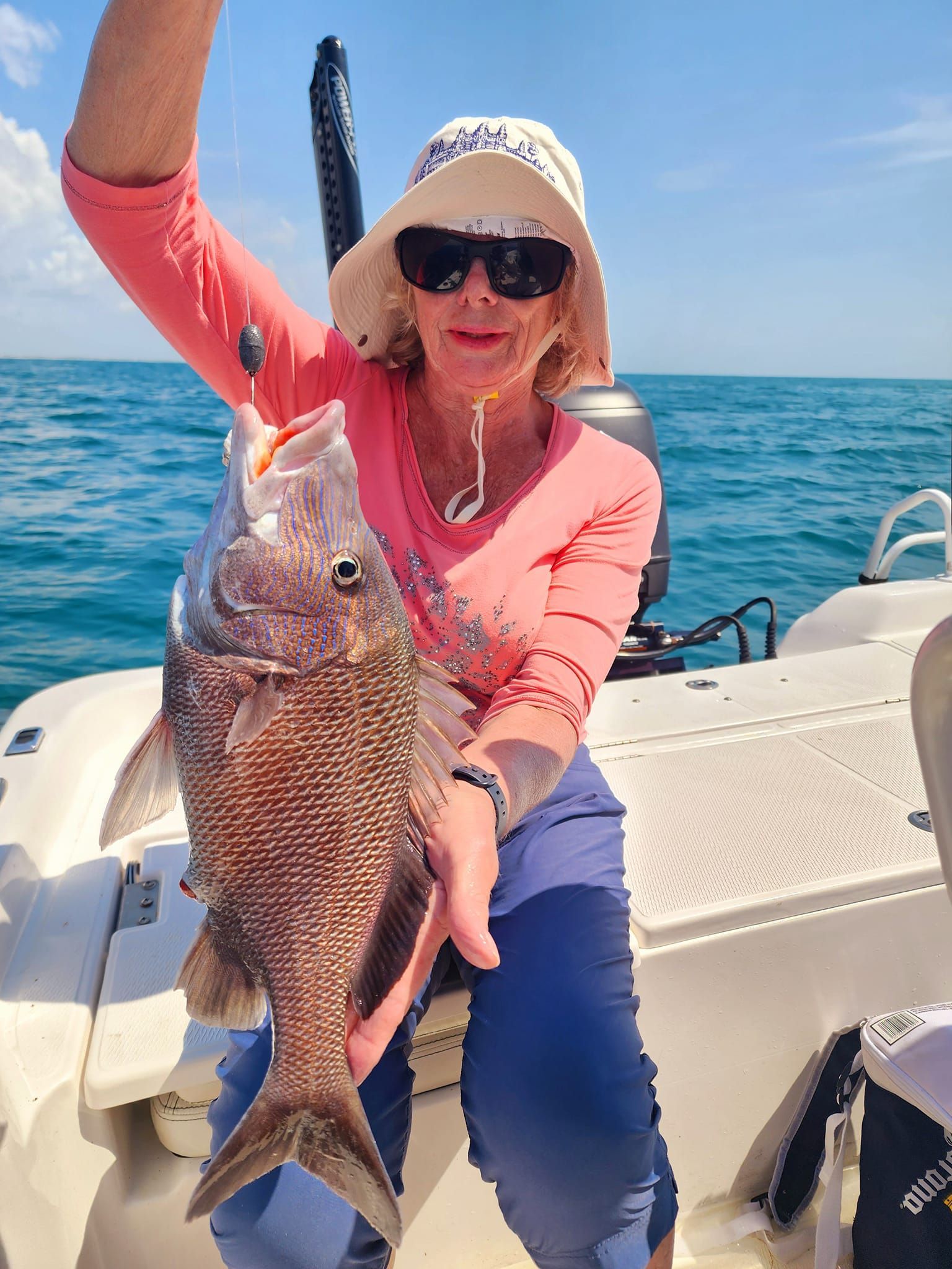 Woman on a boat holds up a freshly caught fish against a sunny, blue ocean backdrop.