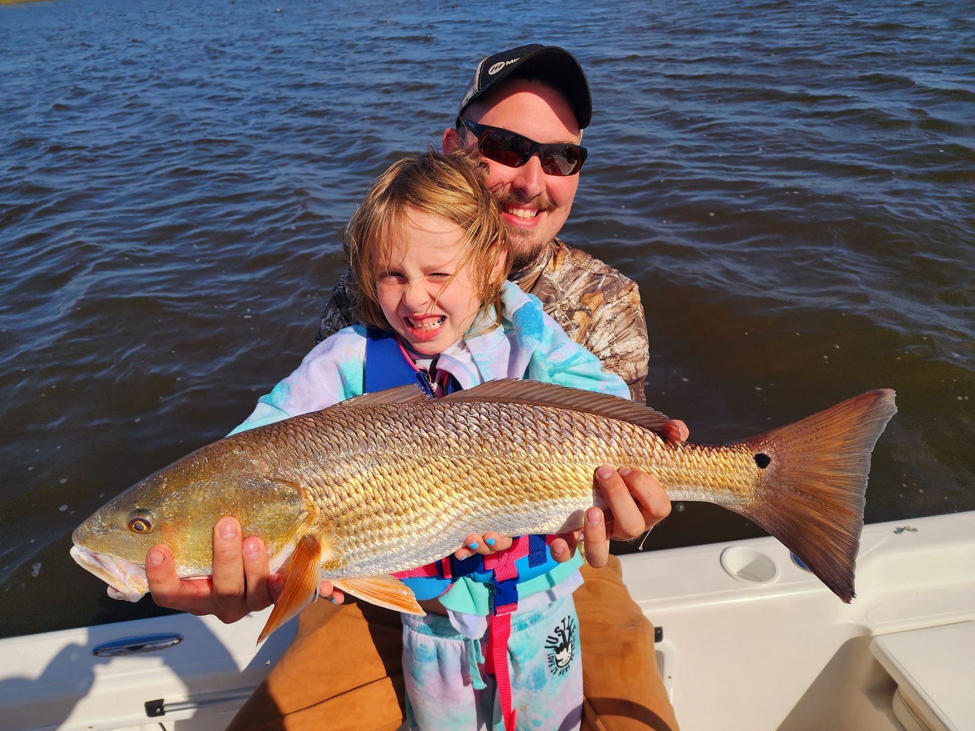 Man and young child smiling, holding a large reddish fish on a boat.