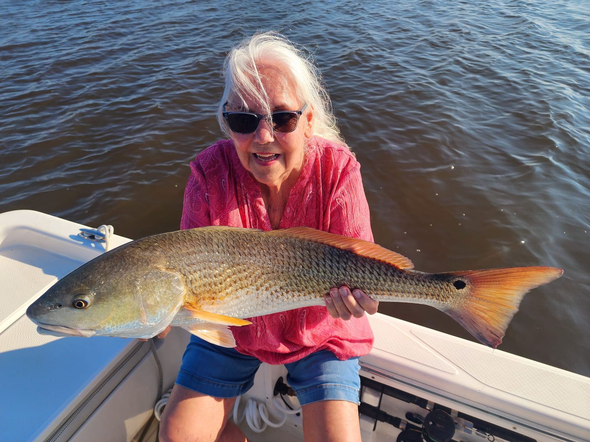 Woman in sunglasses holds a large redfish on a boat, smiling near water.