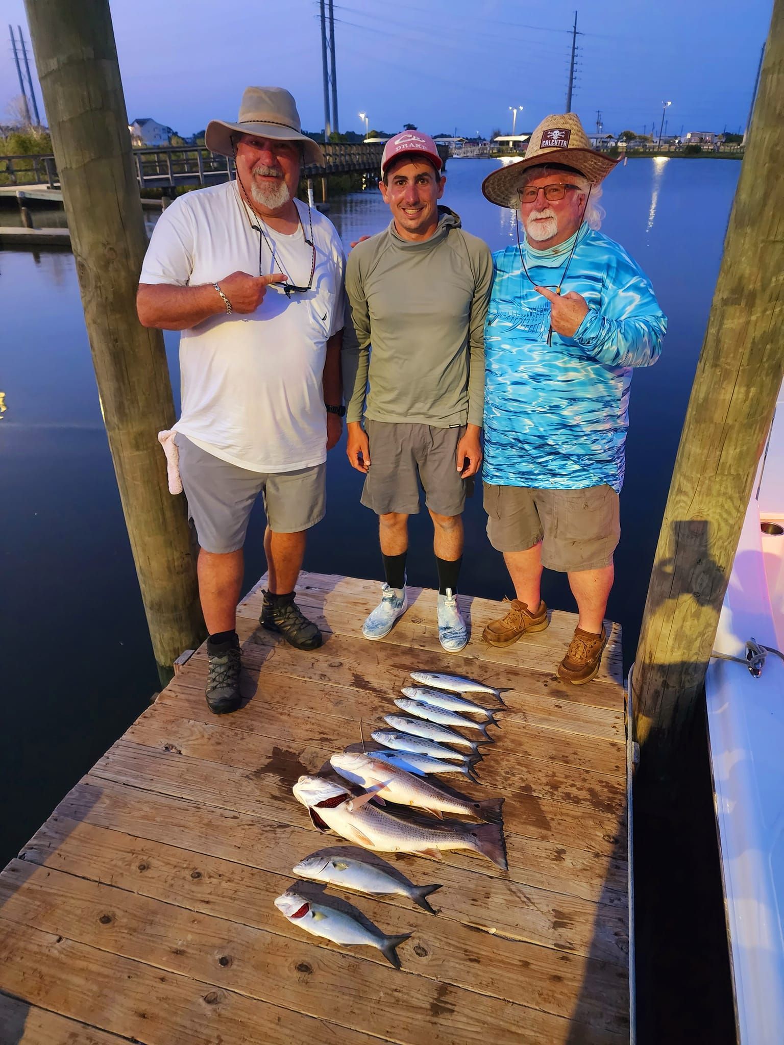 Three men pose with a fish haul on a dock at dusk. One points, others smile. Fish are in a row.