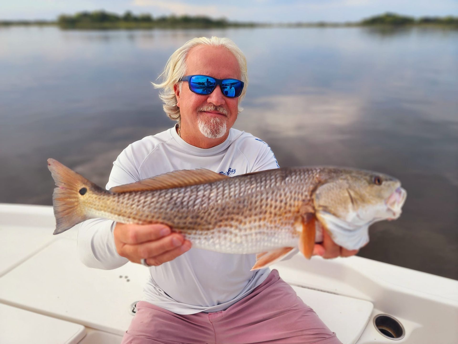 Man on a boat holding a large redfish. Calm water and cloudy sky in background.