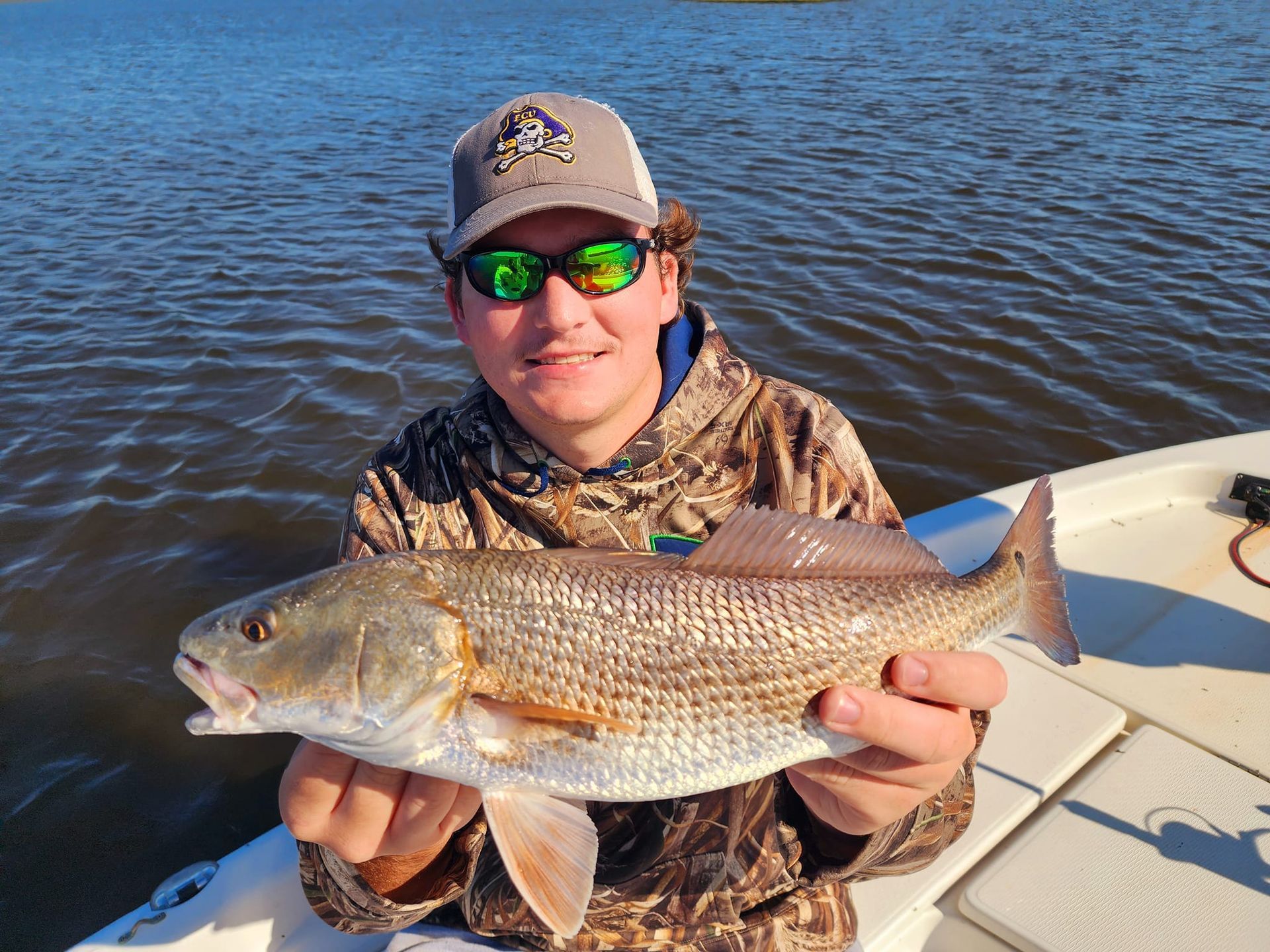 Man holding a reddish fish on a boat, smiling, wearing sunglasses and camouflage jacket. Blue water in background.