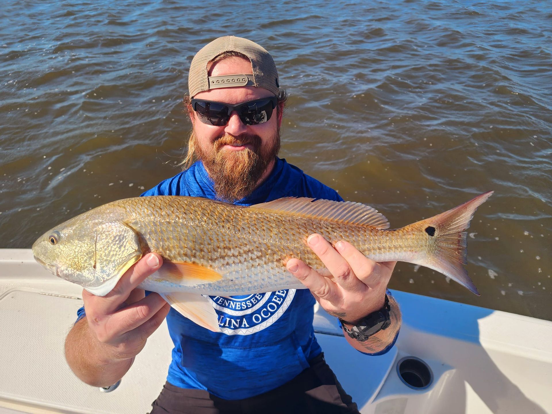 Man holding a redfish on a boat, smiling. He wears sunglasses, a cap, and a blue shirt. The water is brown.