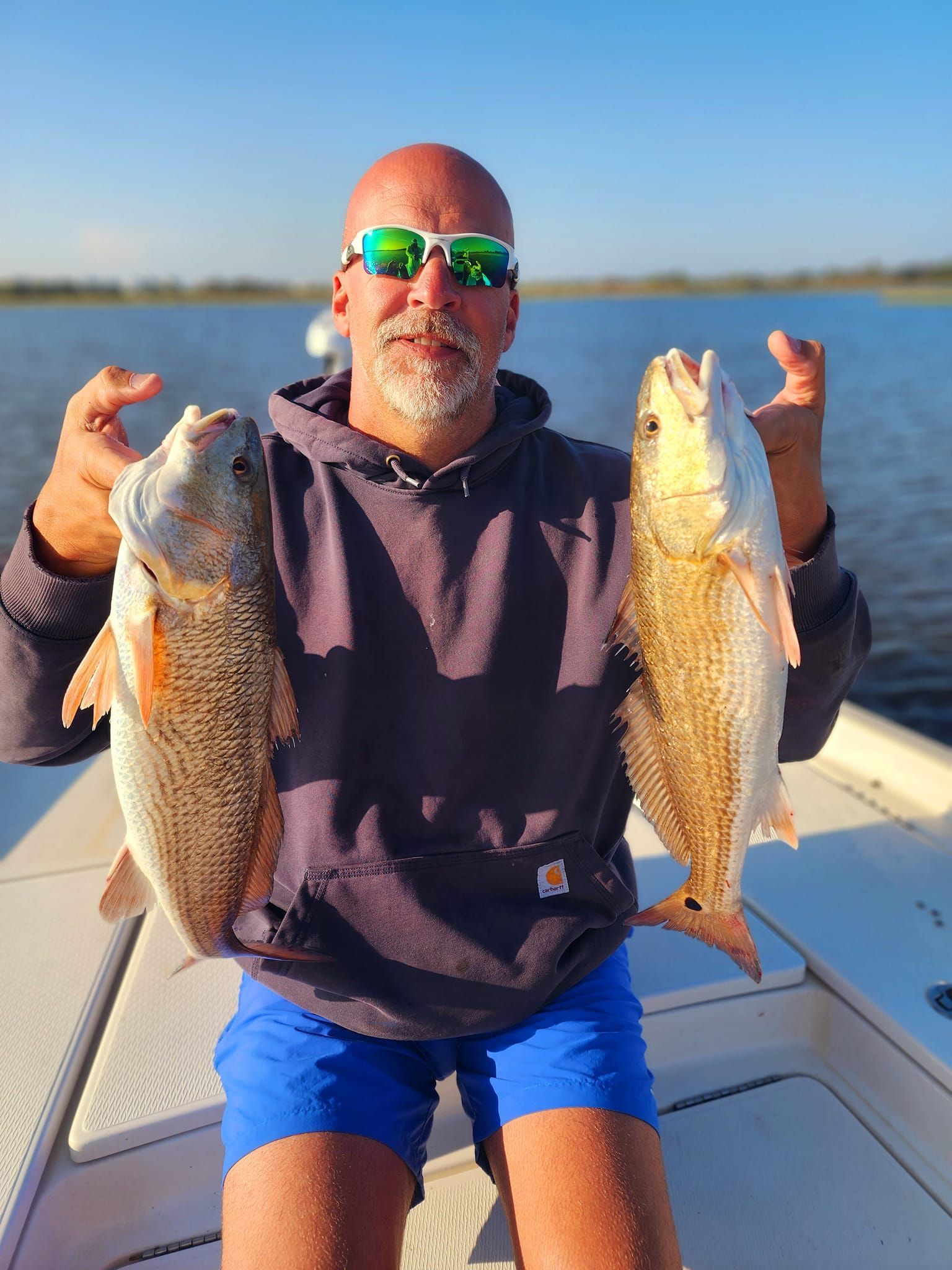 Man holding two redfish on a boat, blue water in the background.