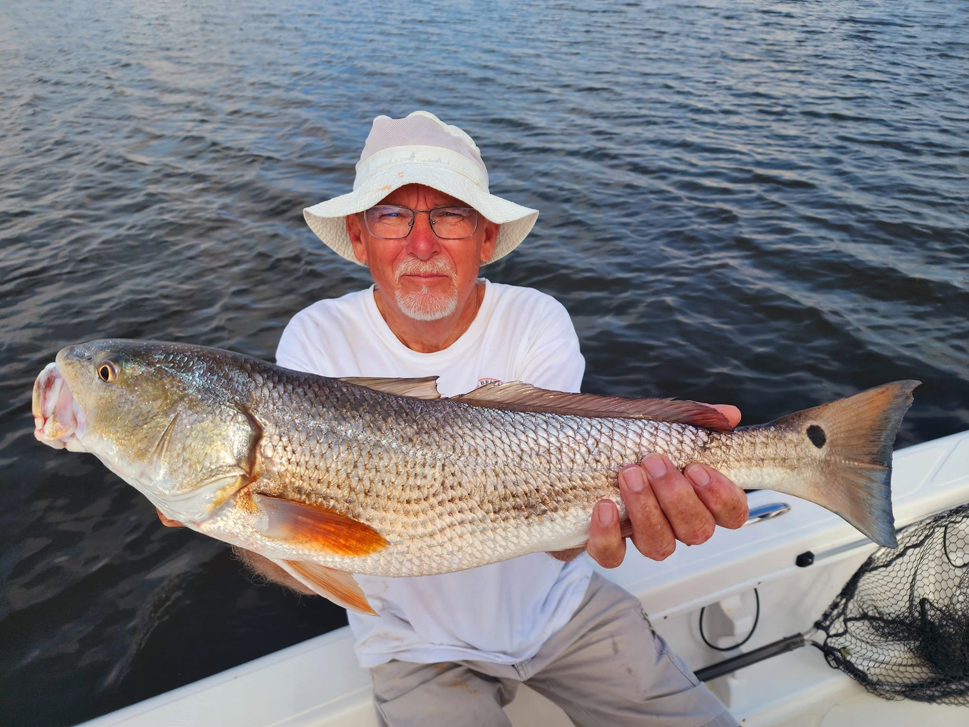 Man in a hat holding a large redfish on a boat, against a water backdrop.