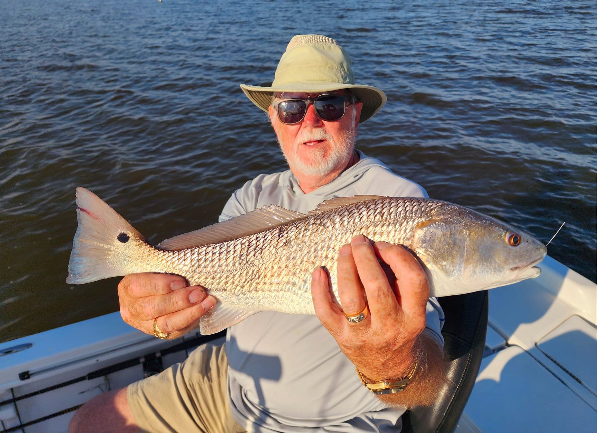 Man in a boat holding a redfish; ocean backdrop; wearing sunglasses and a hat.
