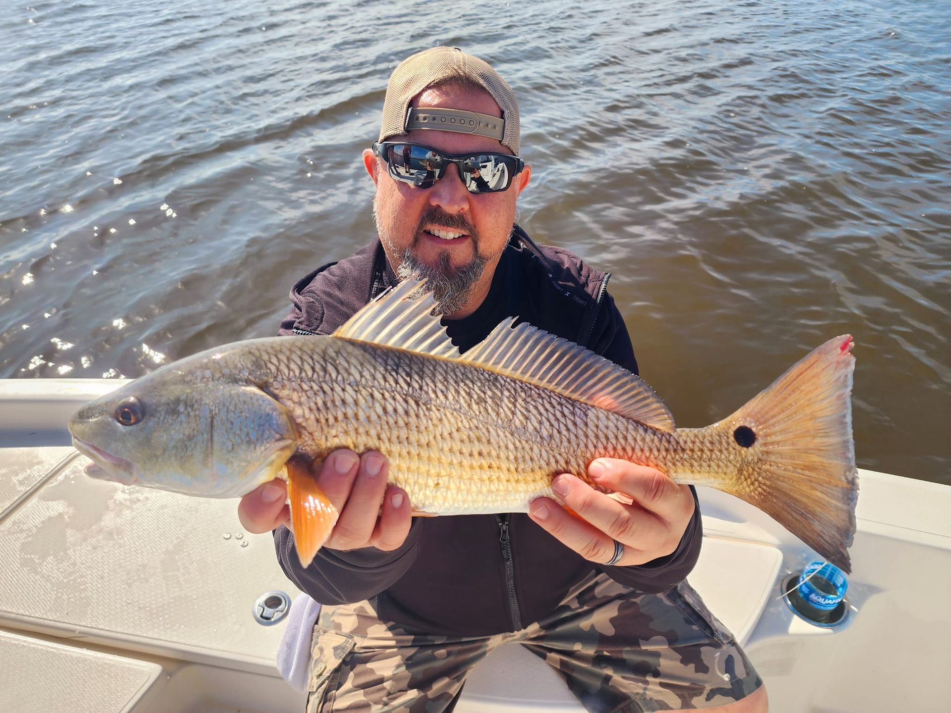 Man on boat holding up a redfish with a red tail, smiling, in sunlight.