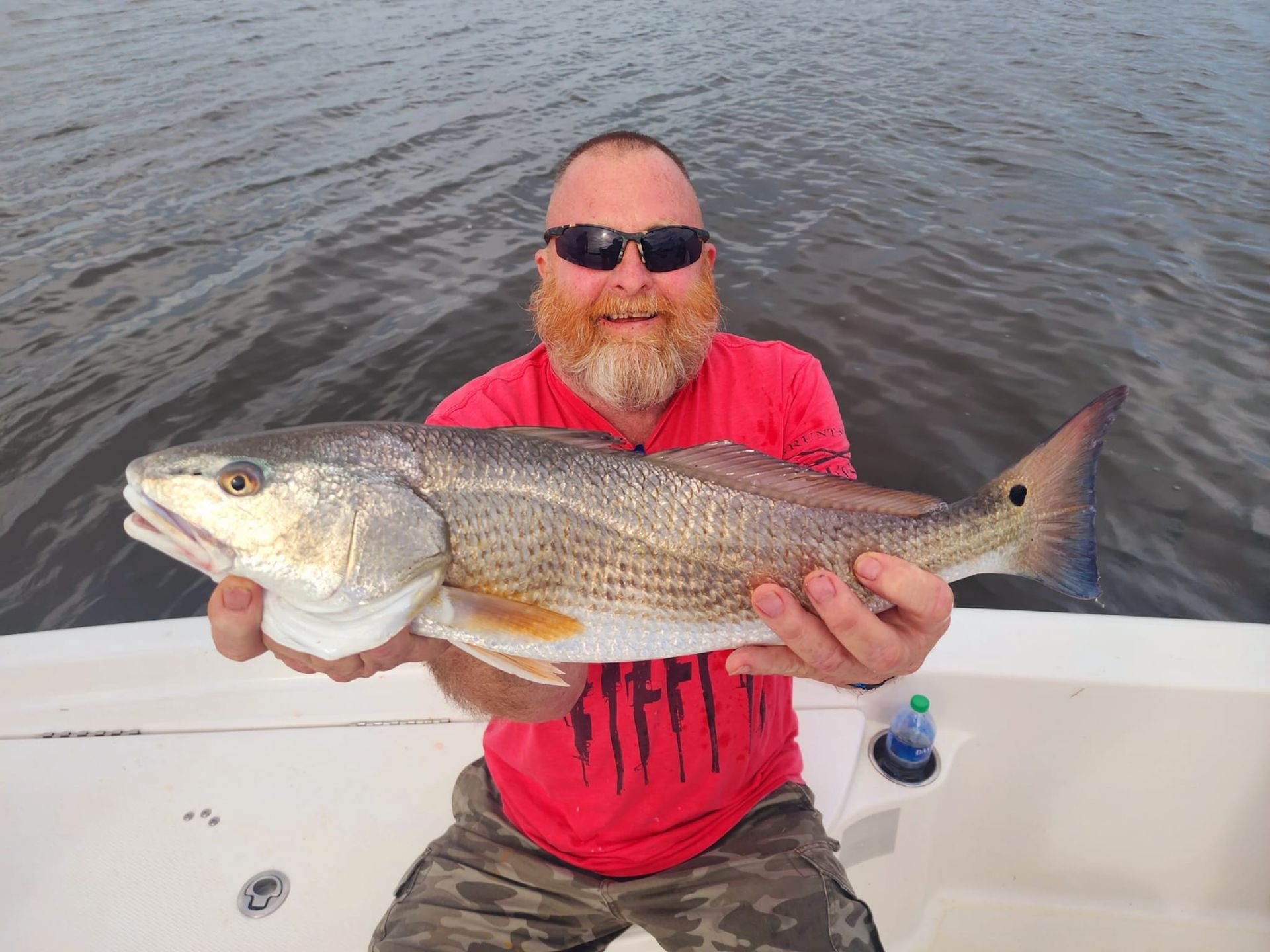 Man holding a large redfish on a boat; water in background. Man has a red beard and sunglasses.