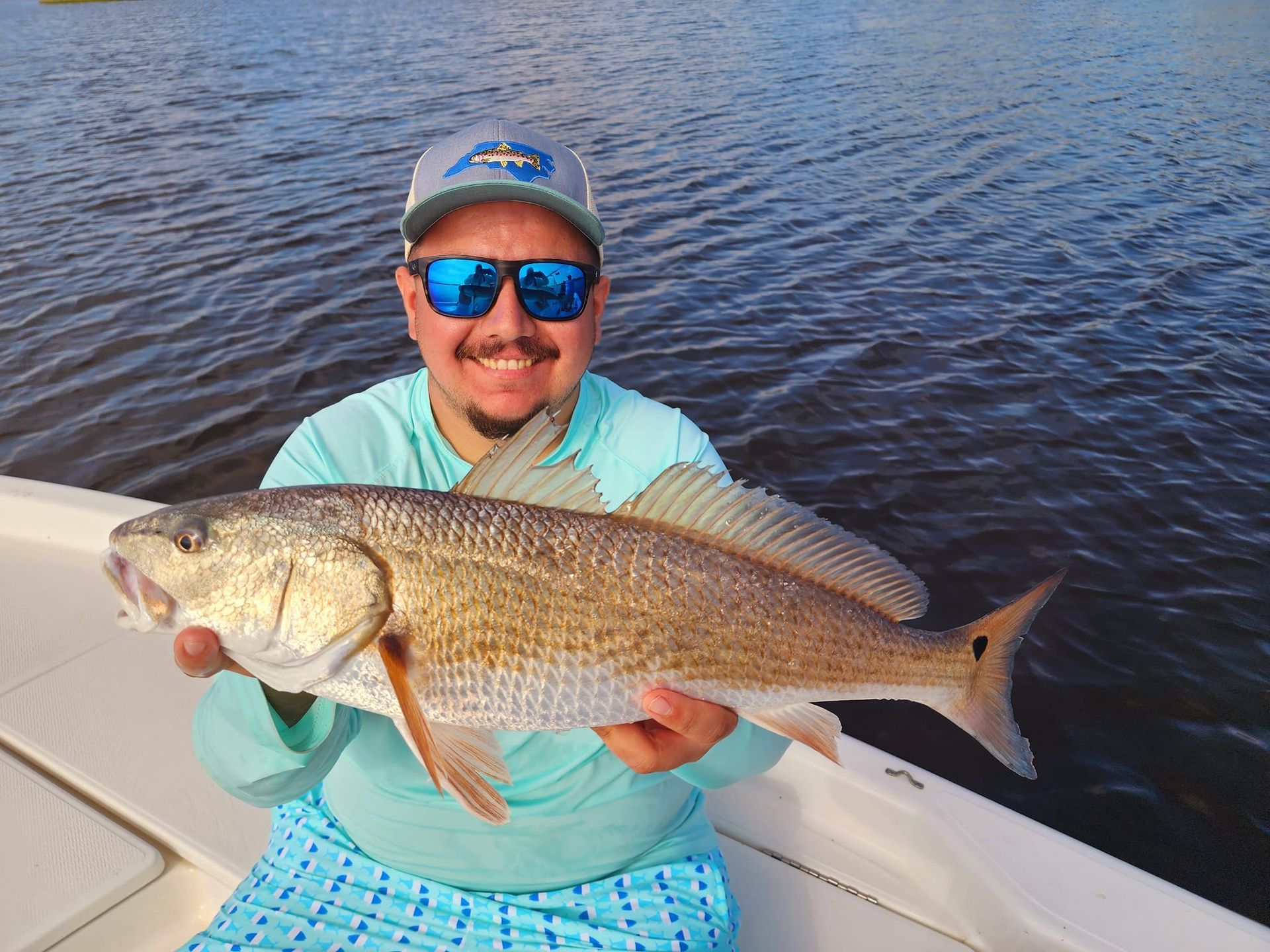Man on a boat holds a large redfish, smiling. The water is visible in the background.