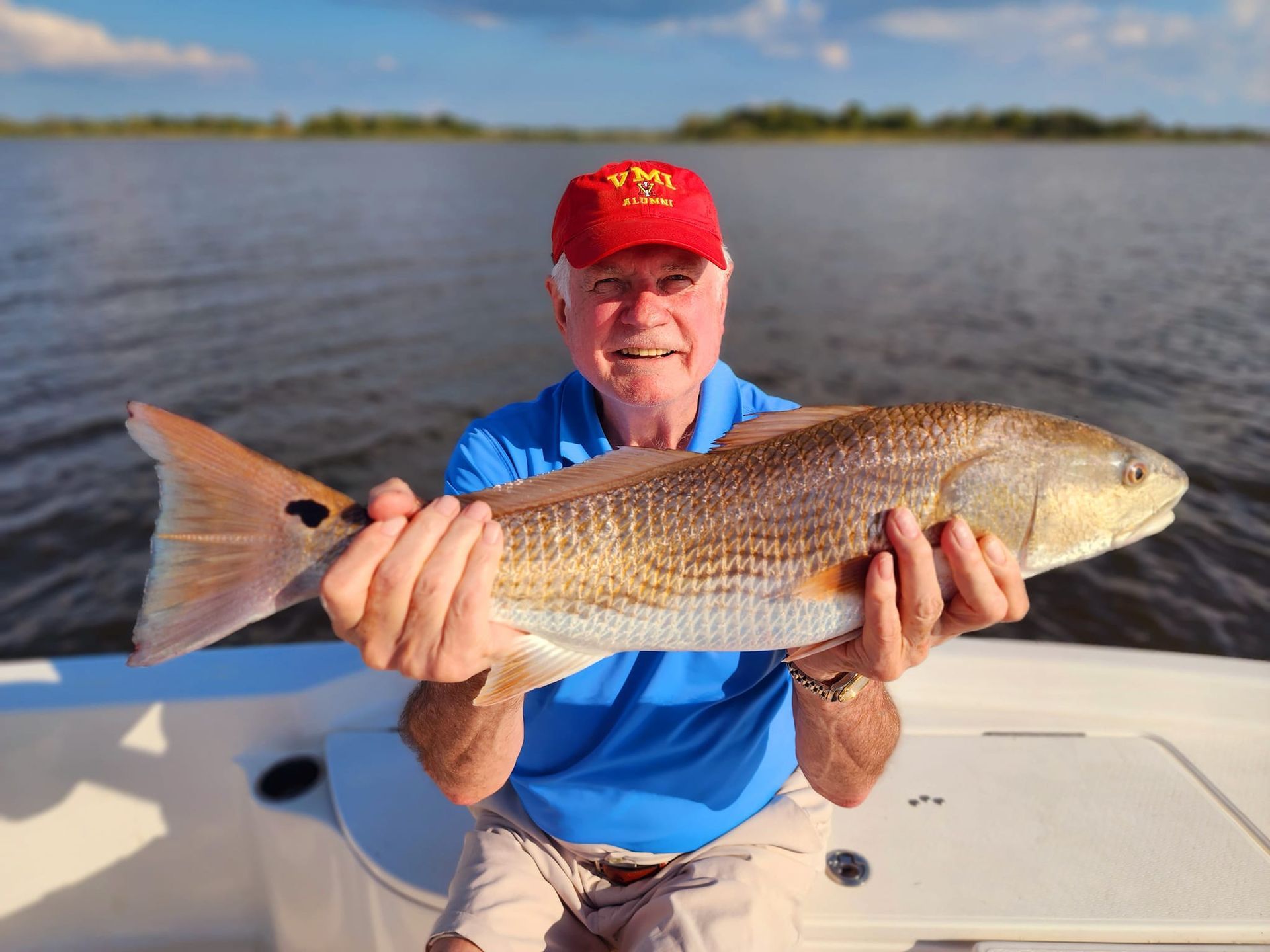 Man holding a large redfish, on a boat, wearing a red hat, blue shirt, and khaki shorts. Water and sky in background.
