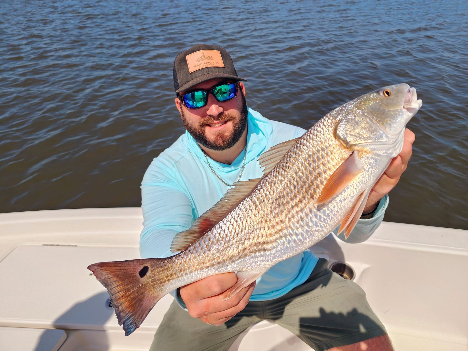 Man holding a large redfish on a boat; water and sunny sky in background.