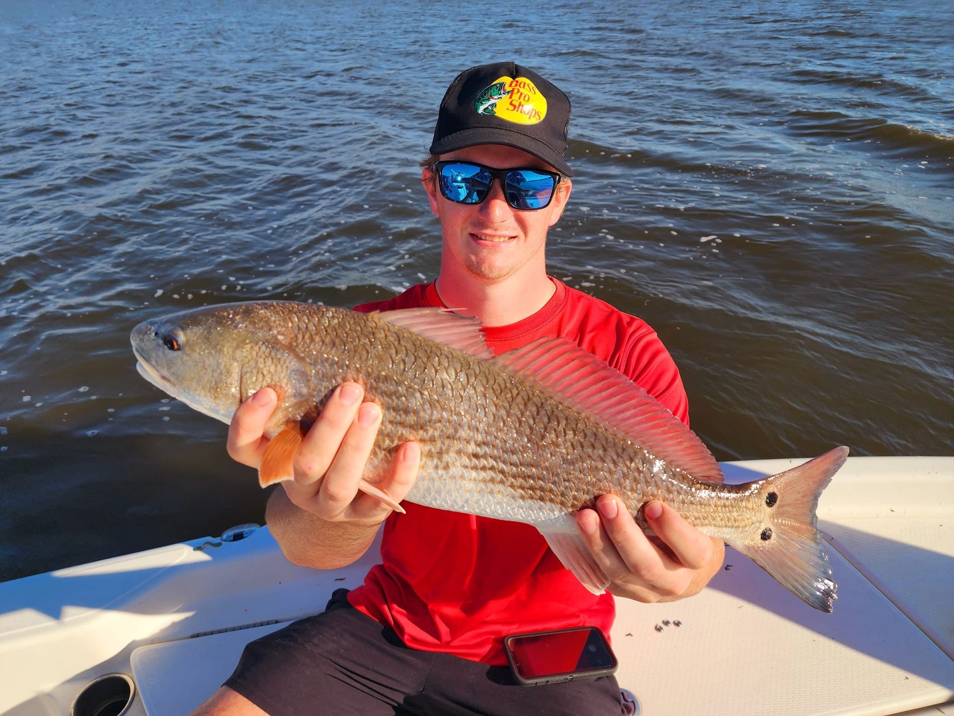 Man holding a redfish on a boat, smiling with the water in the background.
