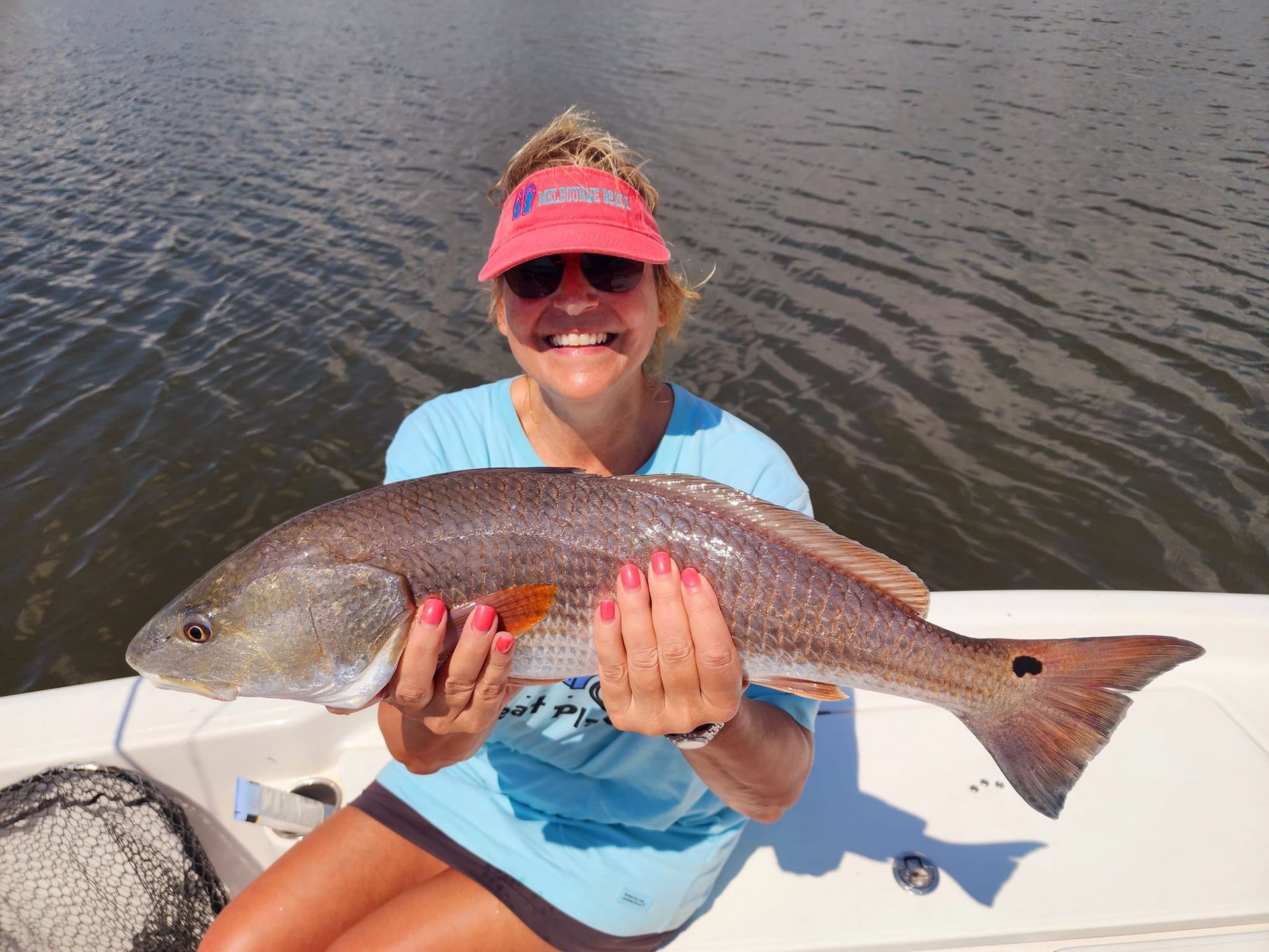 Woman smiling, holding a large redfish on a boat in water, wearing a visor and sunglasses.