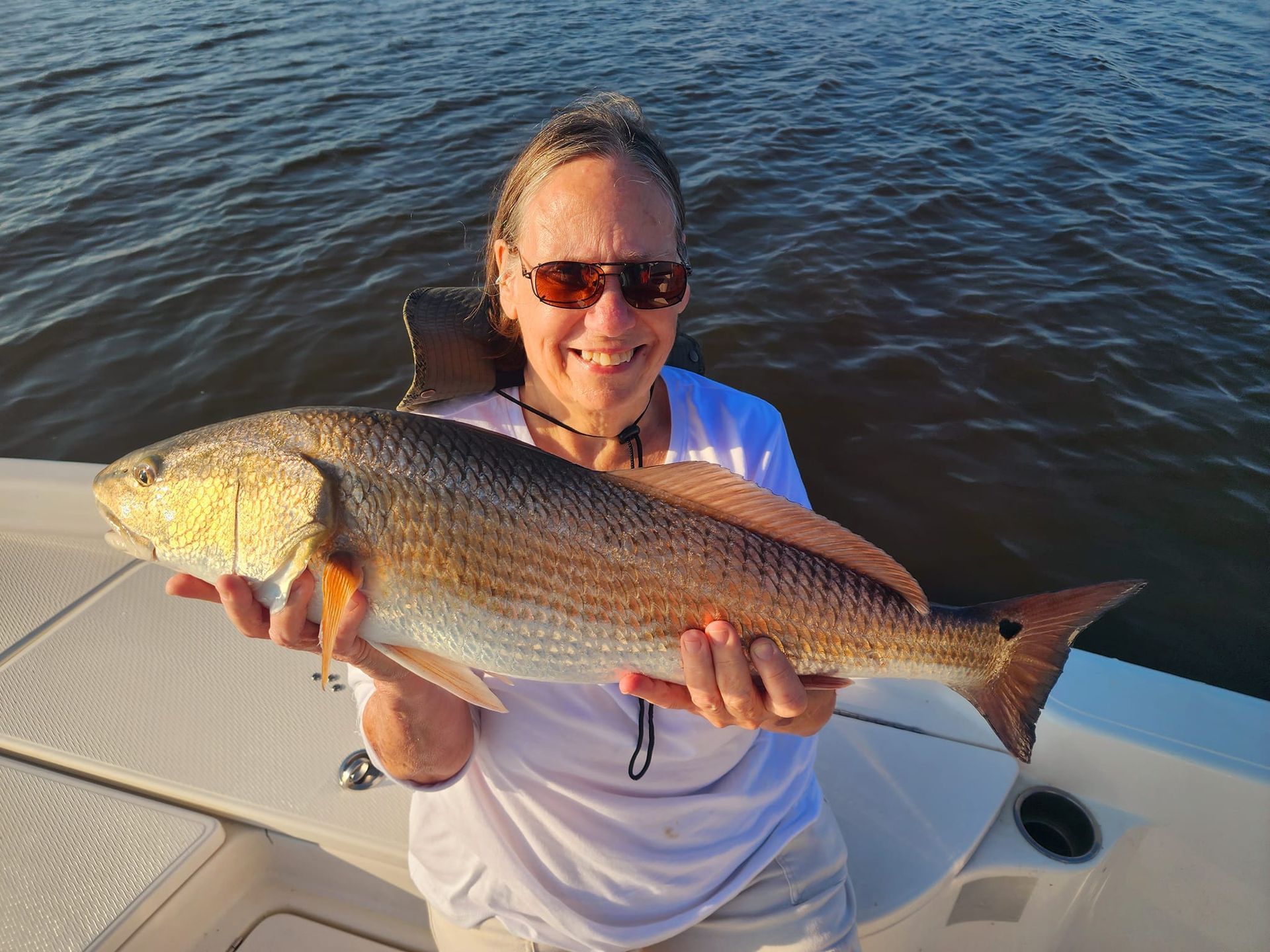 Woman holding a large redfish on a boat; water and setting sun in background.