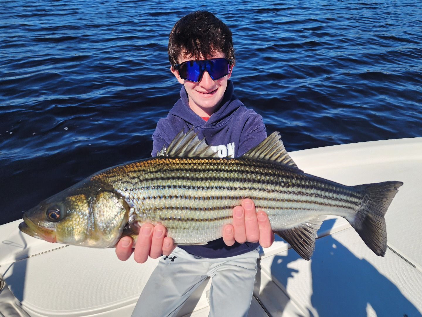 Boy on a boat holds up a striped bass he caught, smiling. Sunny day, dark water.