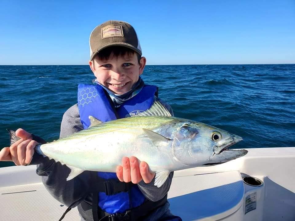 Boy on a boat smiles, holding a freshly caught fish against a blue ocean backdrop.