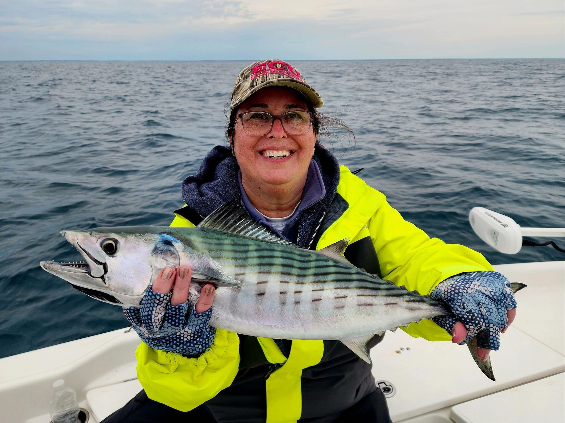 Woman on a boat holding a fish, smiling. She wears a yellow jacket and cap, fishing at sea.