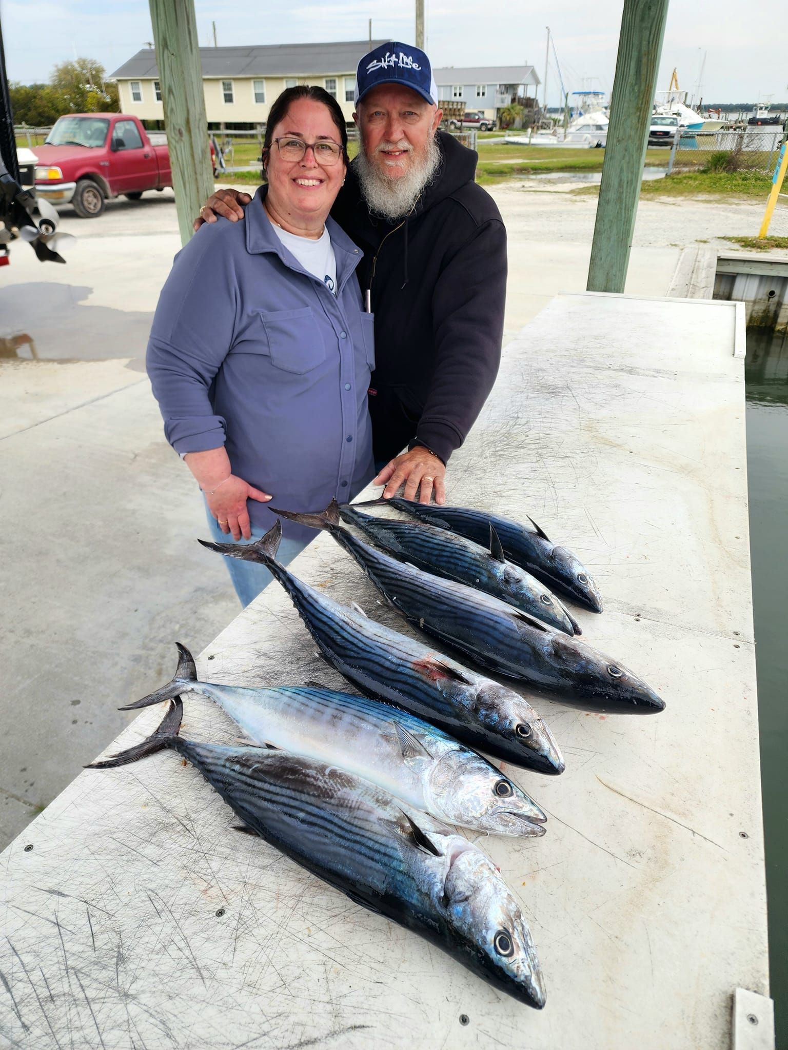 Couple poses with six fish on a dock; woman smiles, man has beard, setting is outdoors, possibly a harbor.