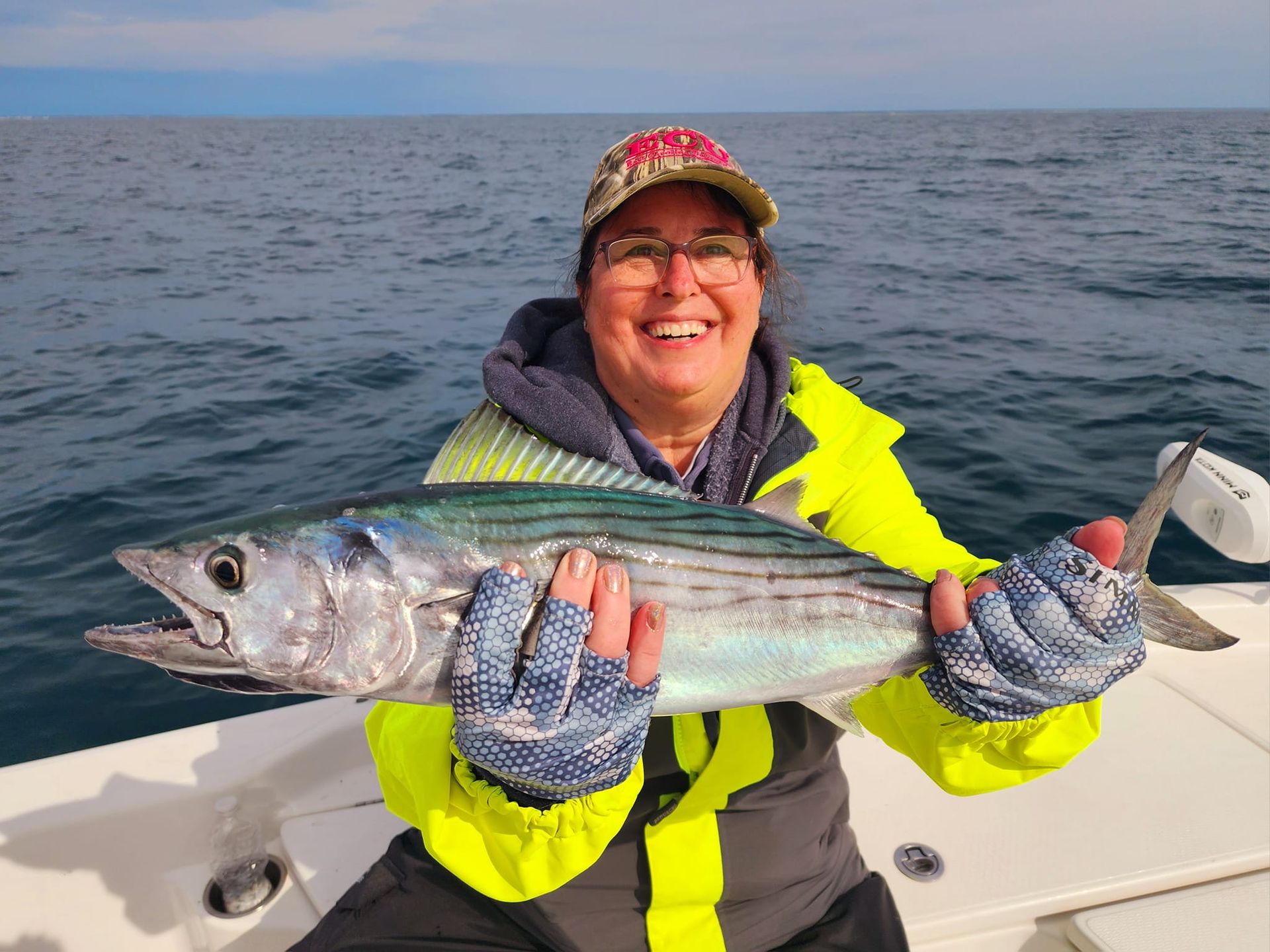 Smiling person holding a fish on a boat, wearing a yellow jacket and hat, blue water in the background.