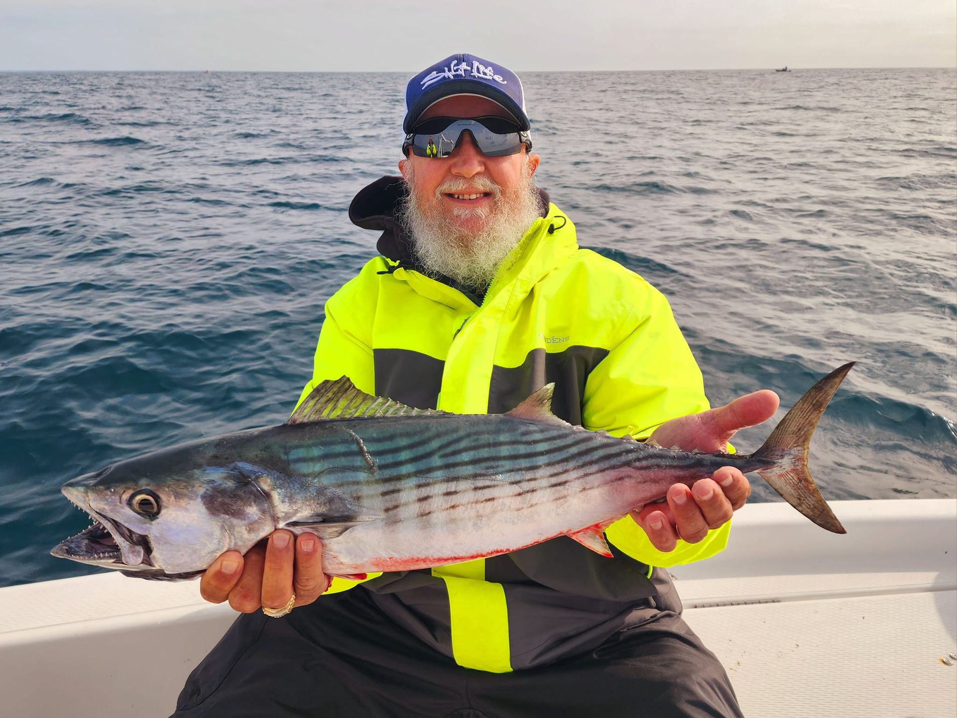 Man holding a striped fish on a boat, wearing a yellow jacket and hat. Ocean in background.