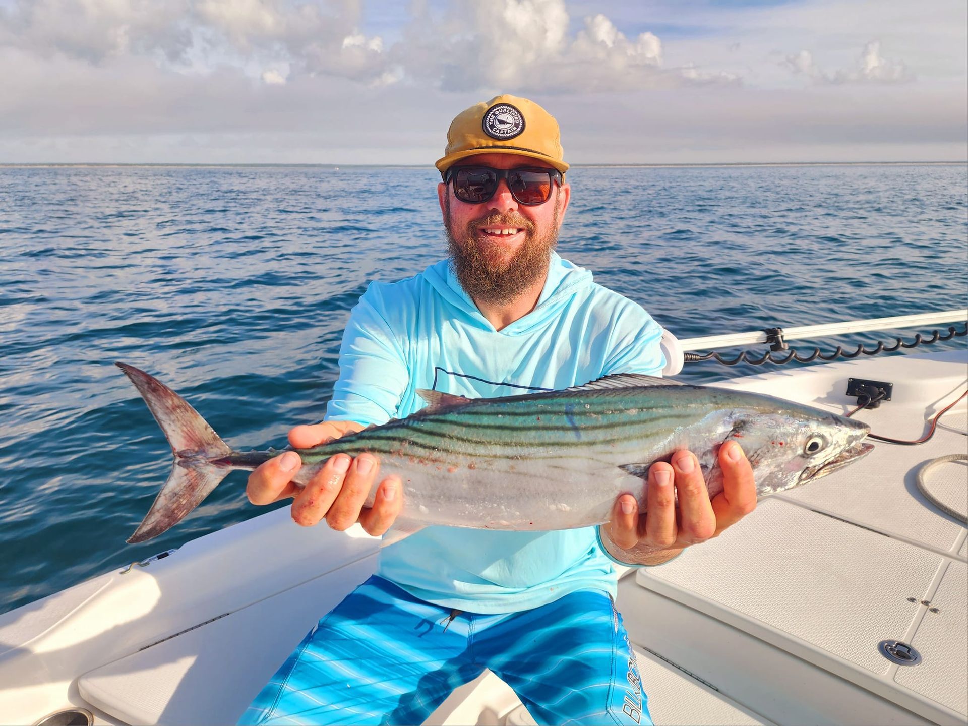 Man holding a fish on a boat; ocean background. He's smiling, wearing sunglasses, and a hat.