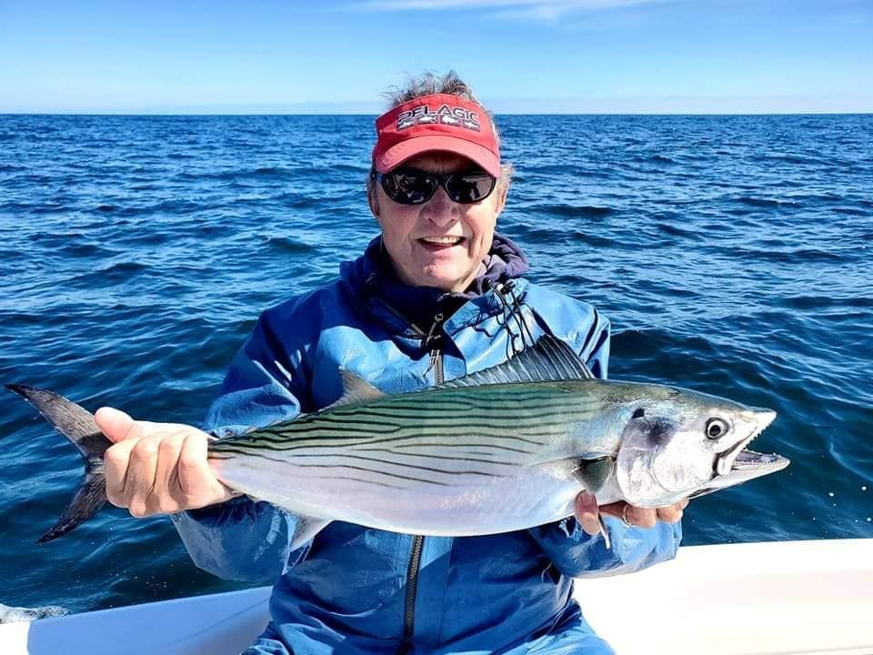 Man holding a fish on a boat; blue jacket, red hat, sunny, ocean.