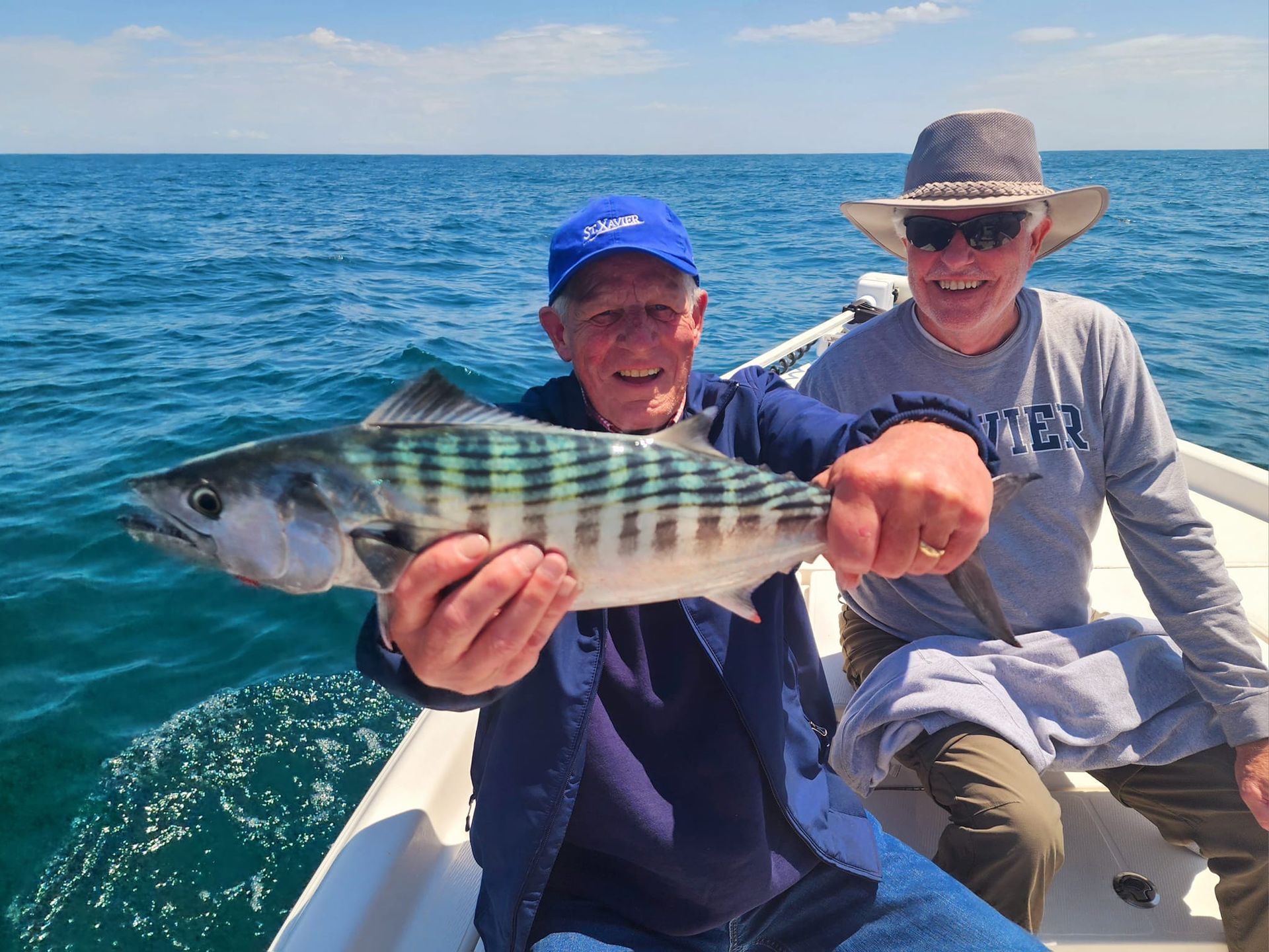 Two men on a boat, one holding a striped fish. They are smiling in the sunlight on the water.