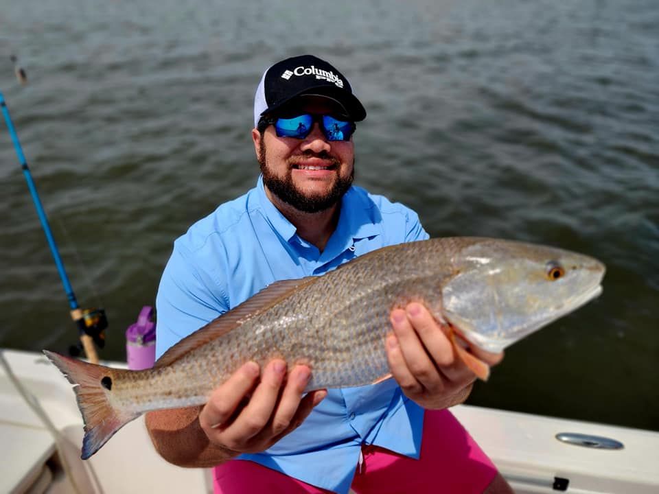 Man on a boat holding a redfish. He's smiling, wearing sunglasses, a hat, and a blue shirt.
