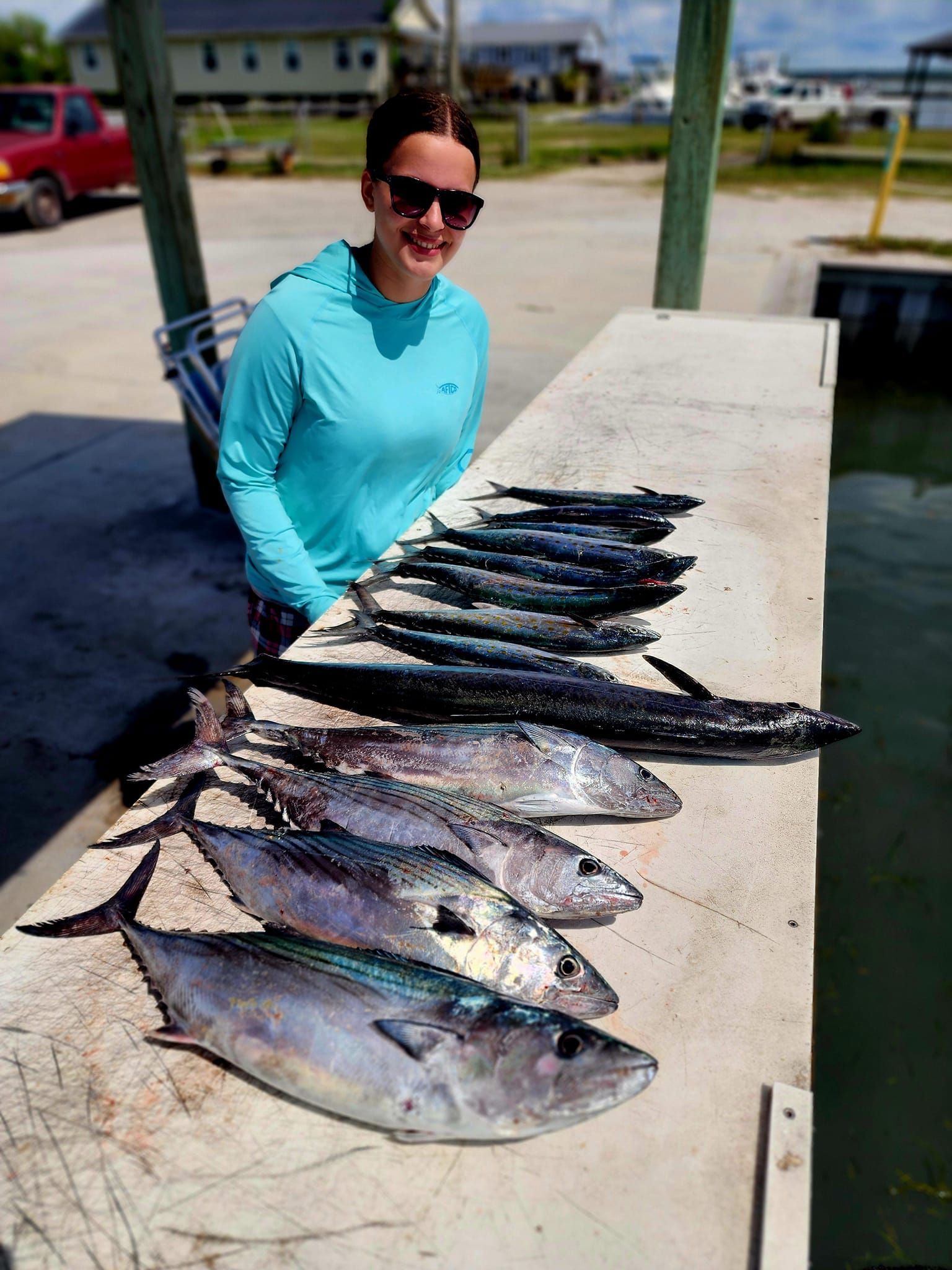 Woman smiling with a large catch of fish on a dock, wearing sunglasses and a blue shirt. Outdoors.