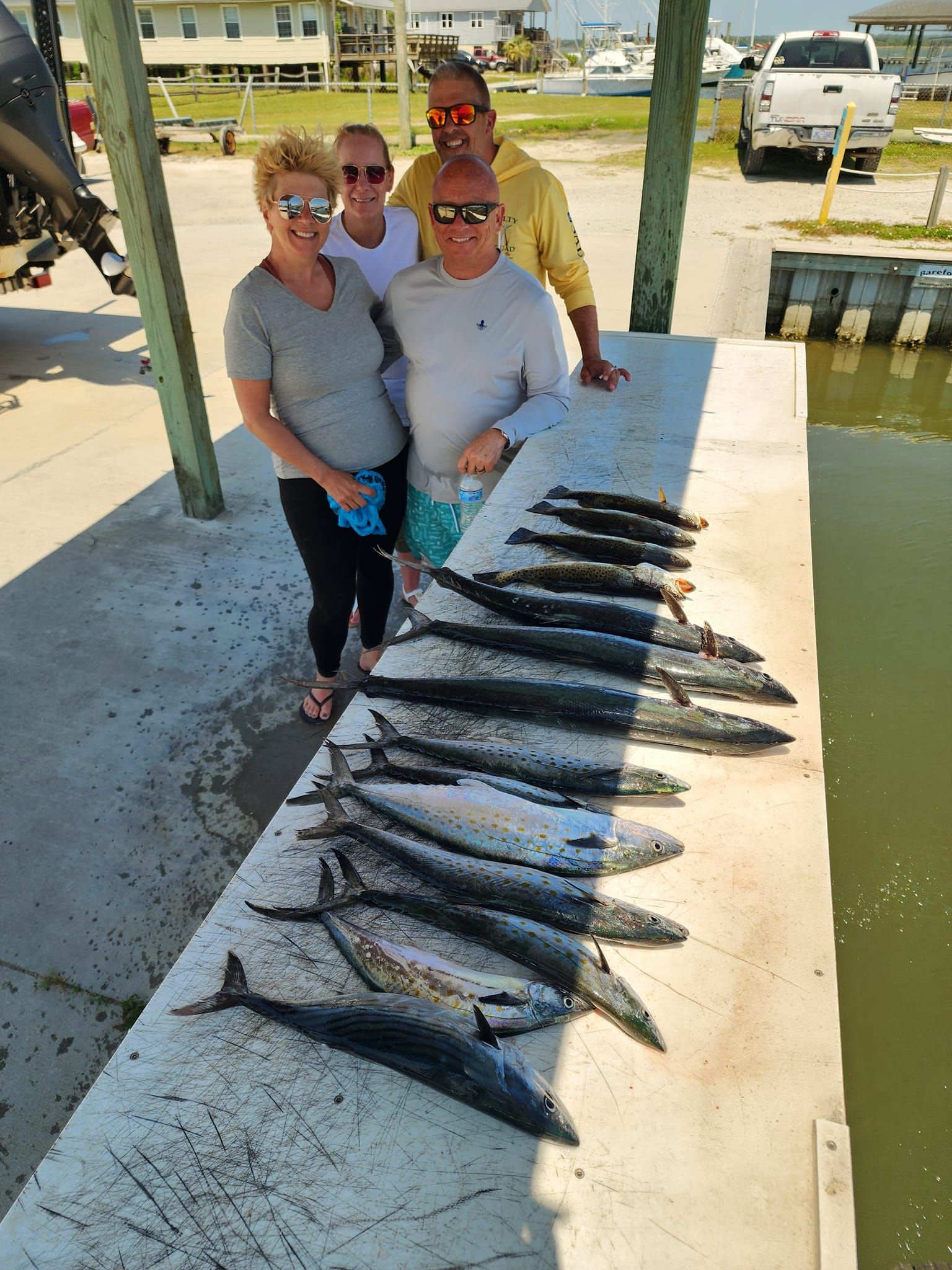 Four people pose with a large catch of fish on a dock, near water.