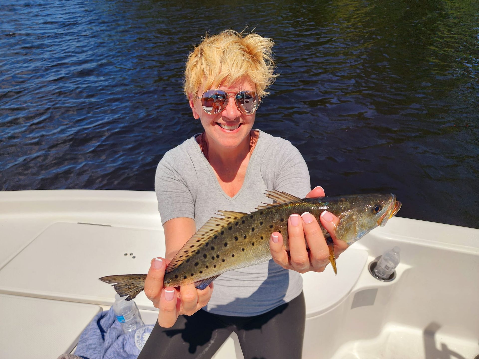 Woman in sunglasses holds spotted fish on boat.