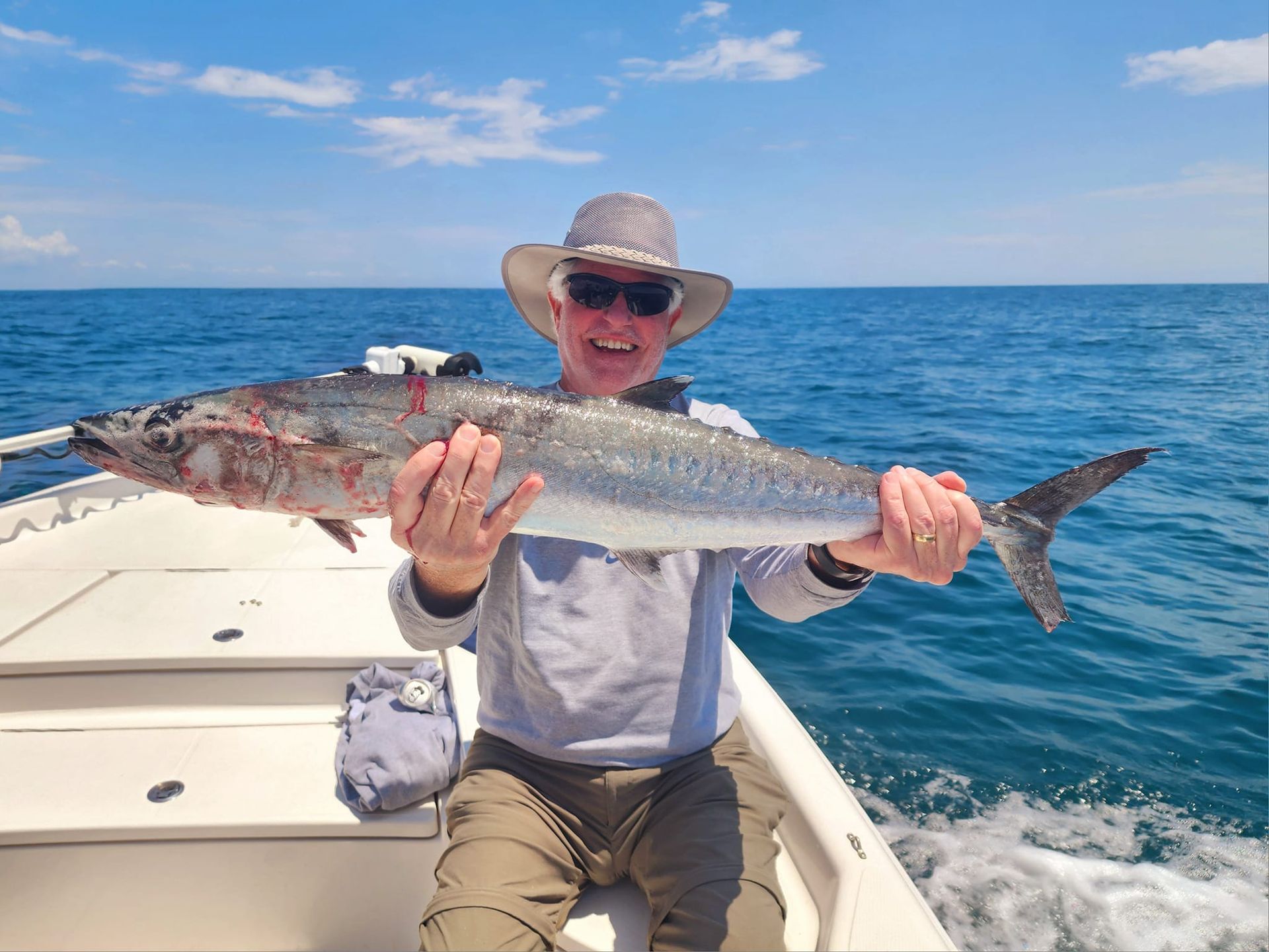 Man on boat holding up a large fish he caught, smiling, blue ocean.