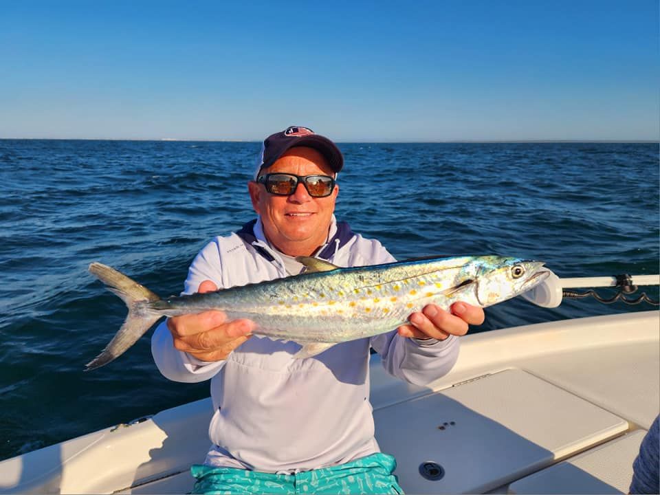 Man on boat holds up a silver fish, ocean backdrop. He wears a hat and sunglasses, smiling.