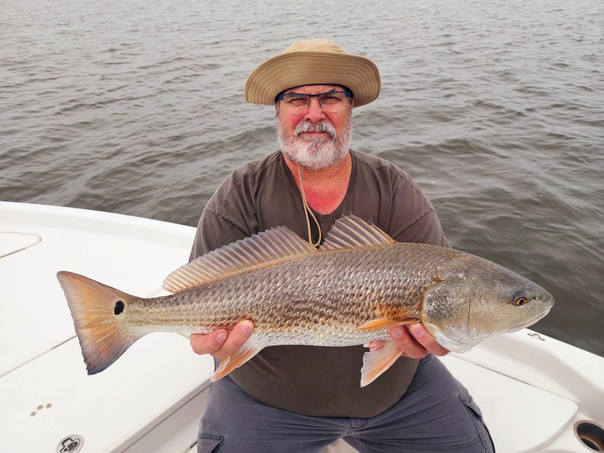Man on boat holding a large red drum fish; outdoors with overcast sky.