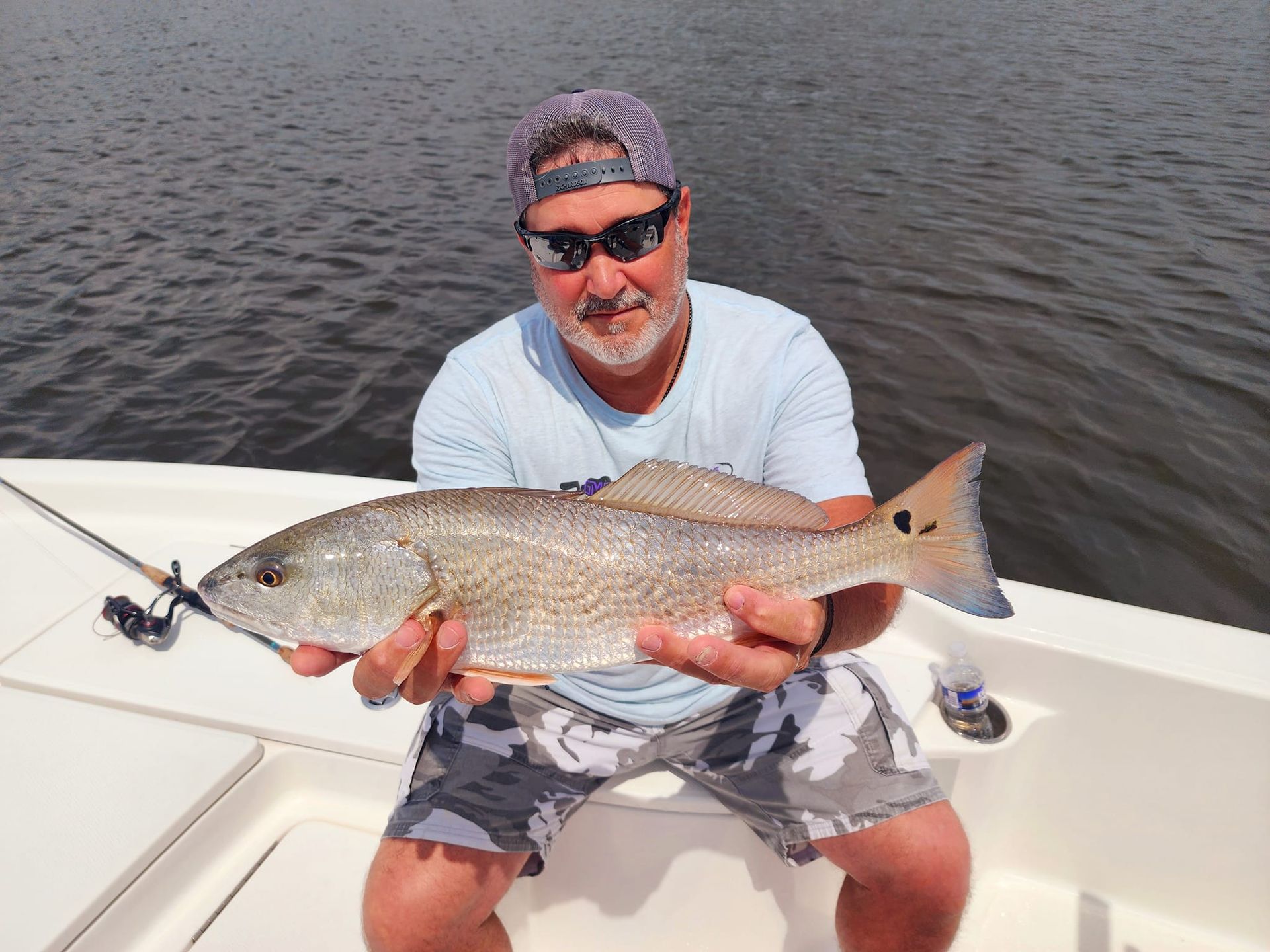 Man on a boat holding a redfish. He is wearing sunglasses and camo shorts.