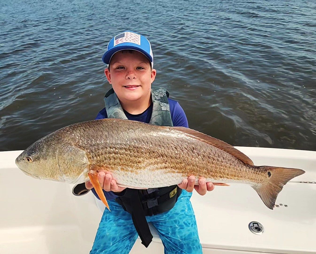 Boy holding large redfish on a boat, smiling. He wears a hat, life vest, and blue pants. Water background.