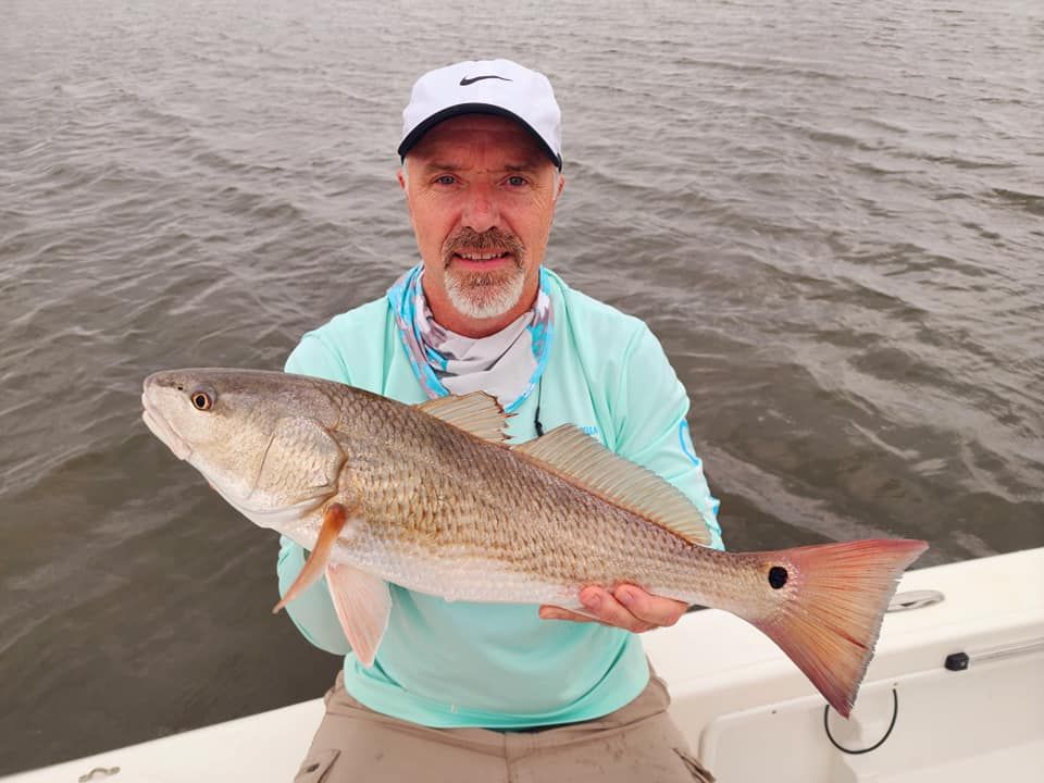 Man on a boat holding a redfish with a red tail. He's wearing a hat, blue shirt, and the background is water.