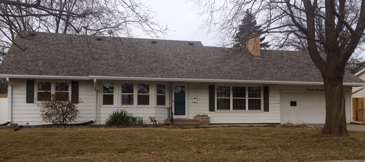 White ranch house with dark shutters, brown roof, and bare trees.