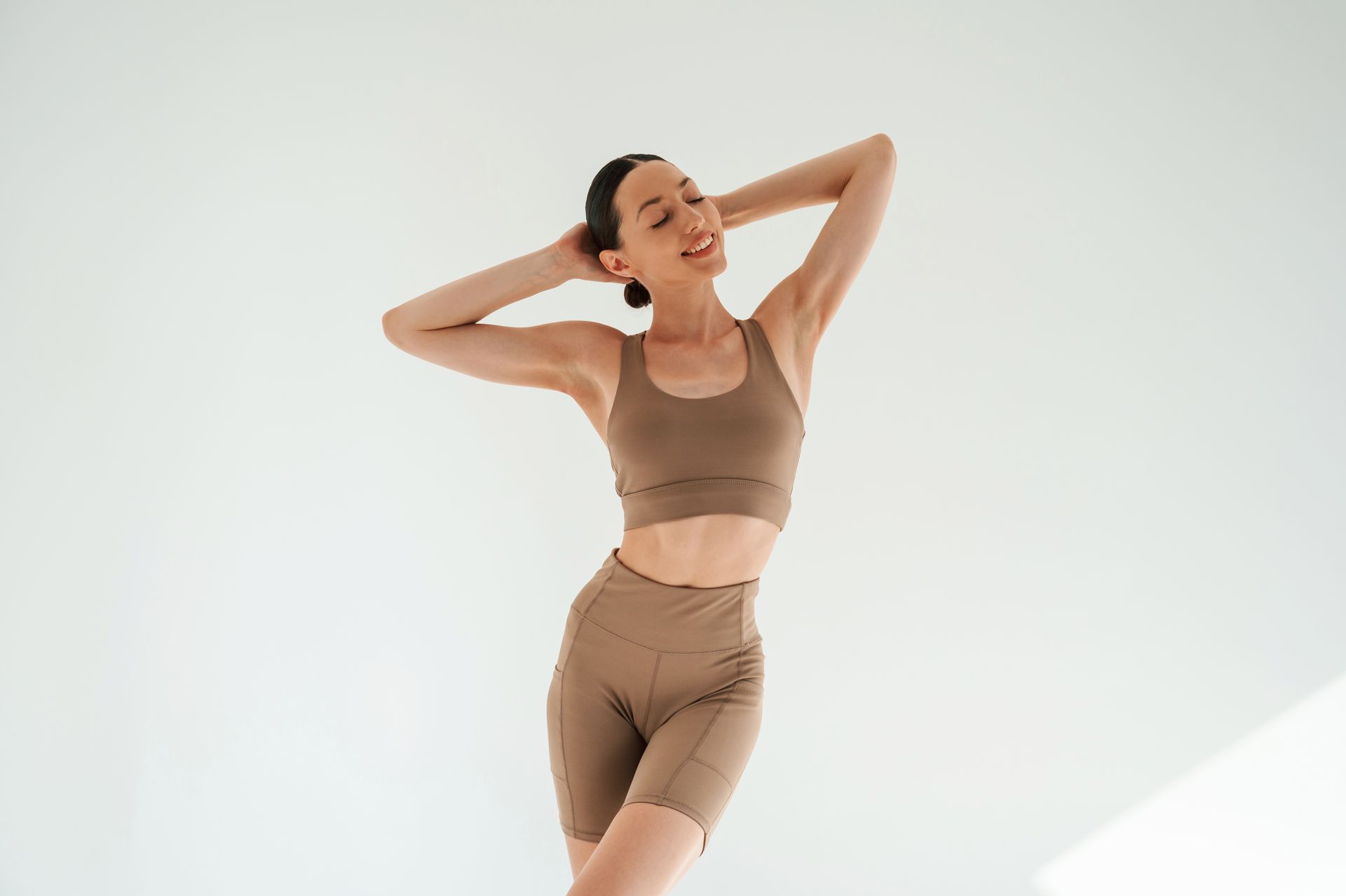 Woman in brown athletic wear, smiling with hands behind head, in a white studio.