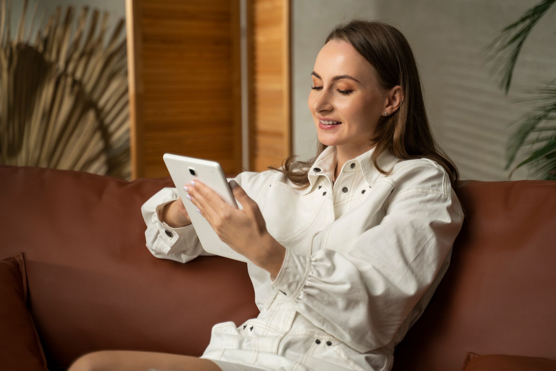 Woman in white outfit sitting on brown couch, holding tablet, smiling.