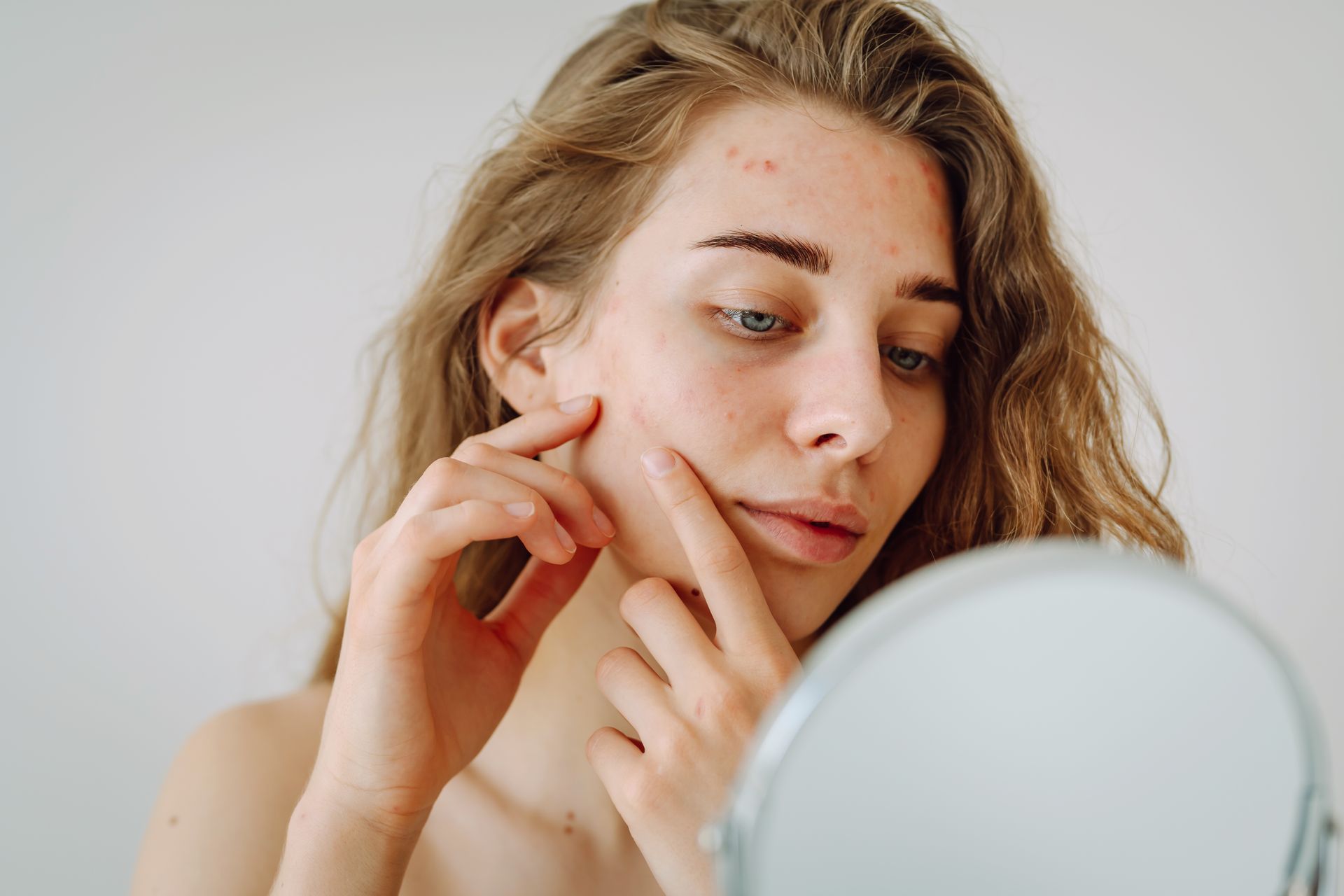 Woman looking at blemishes on her face in a mirror, touching her cheek with fingers, indoors.